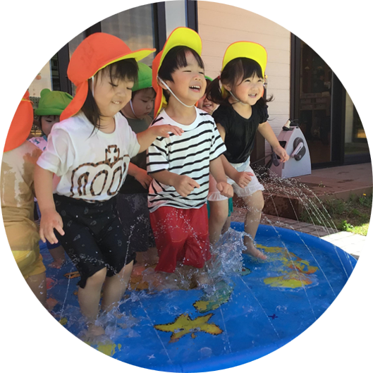 屋外で、複数の幼児が水遊び用のビニールプールで楽しそうに水しぶきを上げている。数人がオレンジや黄色の帽子をかぶっている。
