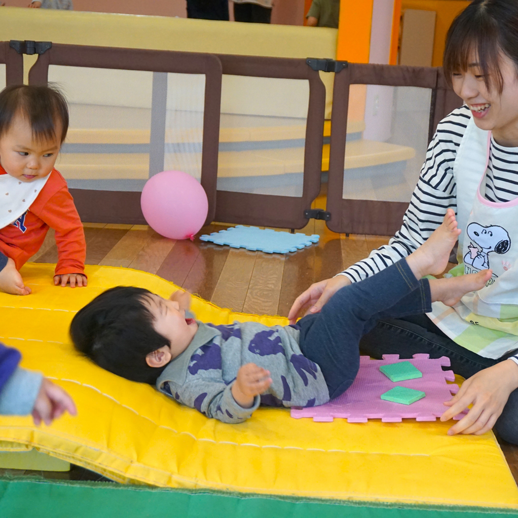 屋内で、保育士が床のマットに横たわる幼児と触れ合っている。別の幼児がそのそばに座っている。ピンクのボールやベビーゲートが見える。